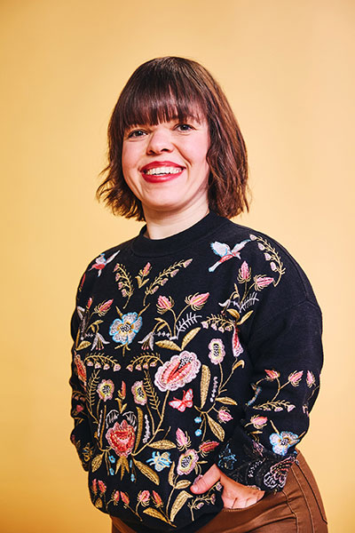Die Fotografin Anna Spindelndreier lächelt in die Kamera. Sie trägt ein buntes Oberteil, hat braune schulterlange Haare und trägt roten Lippenstift.