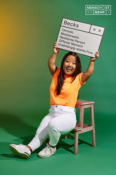 Eine junge Frau mit dunklen Haaren und orangenem Top strahlt in die Kamera. Sie ist kleinwüchsig und hält ein Schild hoch: Becks, Christin, Meisterwerk, Resiliente Person, Offener Mensch, Unabhängig-starke Frau