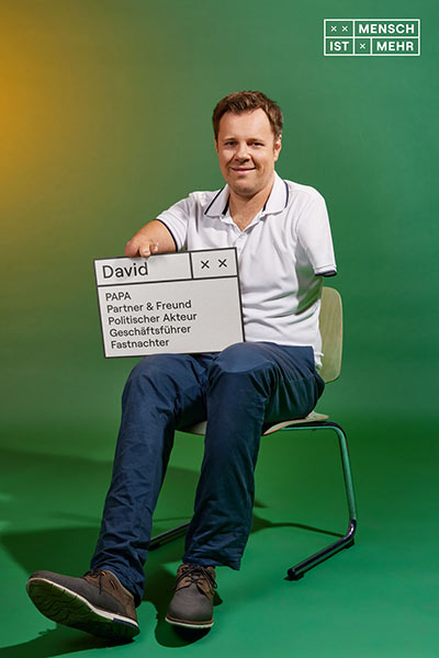 Ein Mann mittleren Alters sitzt lässig auf einem Stuhl und lächelt. Er hat kurze Arme. Auf seinem Schild steht: David, Papa, Partner und Freund, Politischer Akteur, Geschäftsführer, Fastnachter