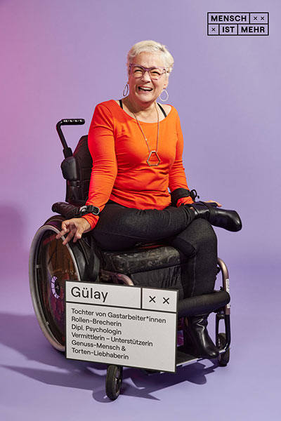 Eine Frau mit orangenem Top sitzt in einem Rollstuhl. Sie lacht herzlich. An ihrem Rollstuhl hängt ein Schild: Gülay: Tochter von Gastarbeiter*innen, Rollen-Brecherin, Dipl.-Psychologin, Vermittlerin, Genussmensch und Tortenliebhaberin