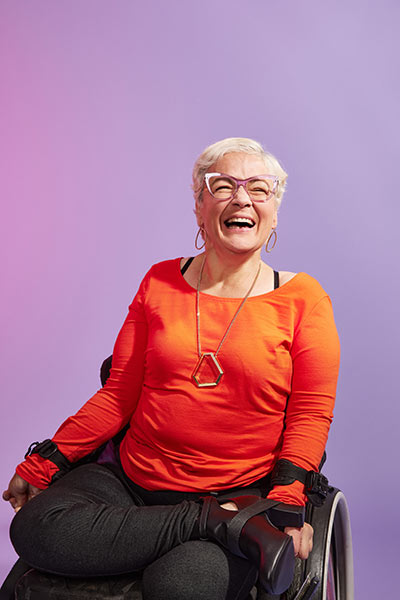 Eine Frau mit weißen Haaren und orangenem Top lacht strahlend der Kamera entgegen. Um ihre Arme liegen Schlaufen, die mit ihrem Rollstuhl verbunden sind. 