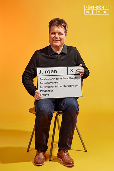 Ein Mann mit dunklem Pullover lächelt und sitzt auf einem Stuhl. Auf seinem Schild steht: Jürgen, Bundesbehindertenbeauftragter, Familienmensch, Jazzmusiker und Literaturliebhaber, Pfadfinder, Freund