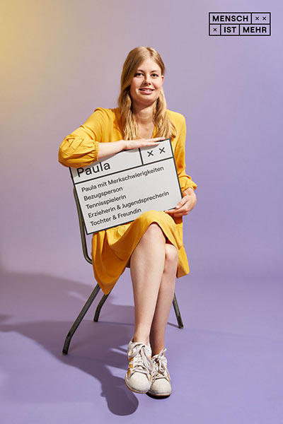 Eine Frau im gelben Kleid lächelt auf einem Stuhl sitzend. Auf ihrem Schild steht: Paula mit Merkschwierigkeiten, Bezugsperson, Tennisspielerin, Erzieherin und Jugendsprecherin, Tochter und Freundin