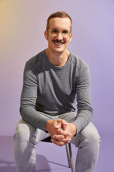 Ein Mann mit grauem Shirt und Schnauzer lacht freundlich.