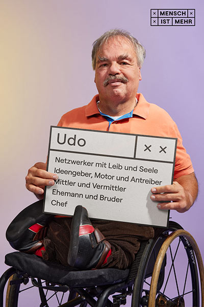 Ein älterer Herr mit Schnauzer lächelt. im Rollstuhl sitzend hält er ein Schild: Udo, Netzwerker mit Leib und Seele, Ideengeber, Motor und Antreiber, Mittler und Vermittler, Ehemann und Bruder, Chef