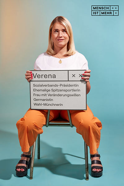 Eine Frau mit blondem Haar schaut sitzend nach oben und lächelt. Sie hält ein Schild: Verena, Sozialverbands-Präsidentin, ehemalige Spitzensportlerin, Frau mit Veränderungswillen, Germanistin, Wahl-Münchnerin